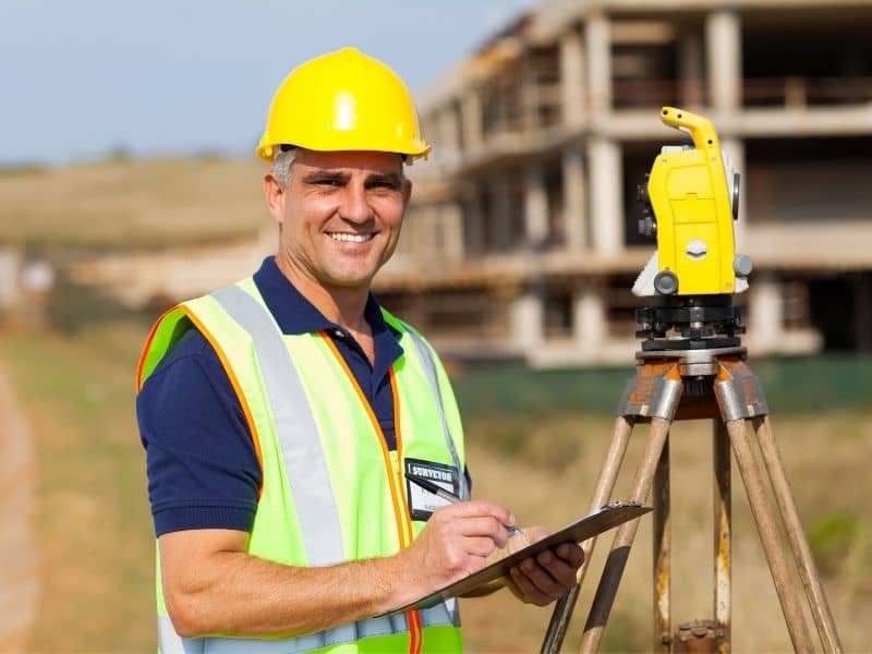 Land surveyor and property owner reviewing undeveloped land near expanding neighborhoods, new roads, and commercial development for future planning.